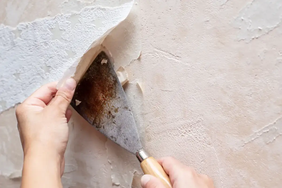 Ręka zeskrobuje starą tapetę szpachlą. Dowiedz się, jak odkleić tapetę ze ściany, by przygotować ją do remontu.