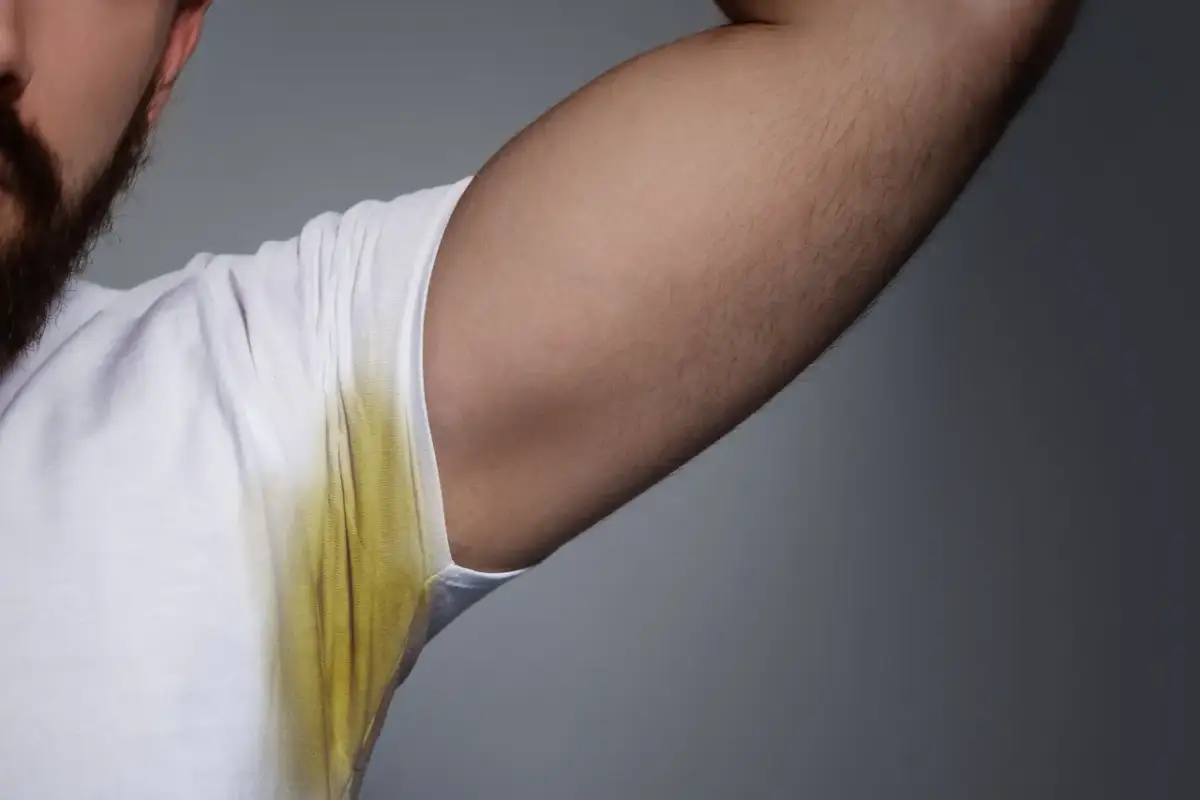 Mężczyzna z zarostem ma białą koszulkę z widocznymi żółtymi plamami pod pachą, świadczącymi o poceniu się.