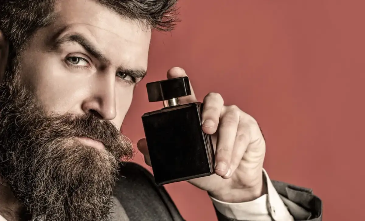 Mężczyzna z brodą trzyma czarną buteleczkę perfum na czerwonym tle.