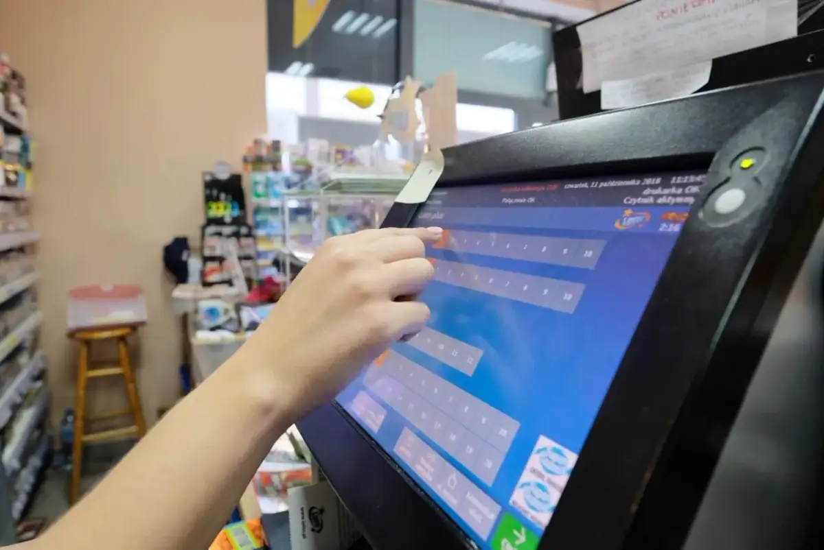 Ręka wybiera liczby na ekranie terminala, pokazując, jak grać w Eurojackpot przez internet.