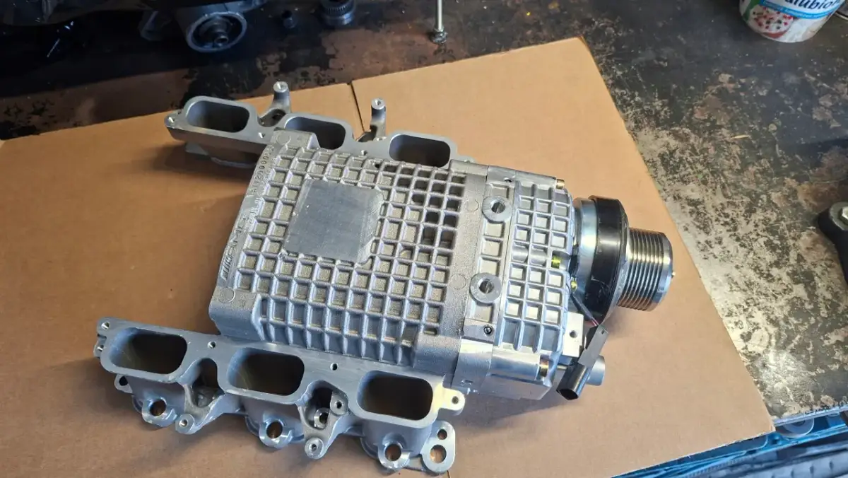 Srebrna sprężarka mechaniczna z kolektorem dolotowym, część do Mercedes Benz C32 AMG, leży na kartonie.