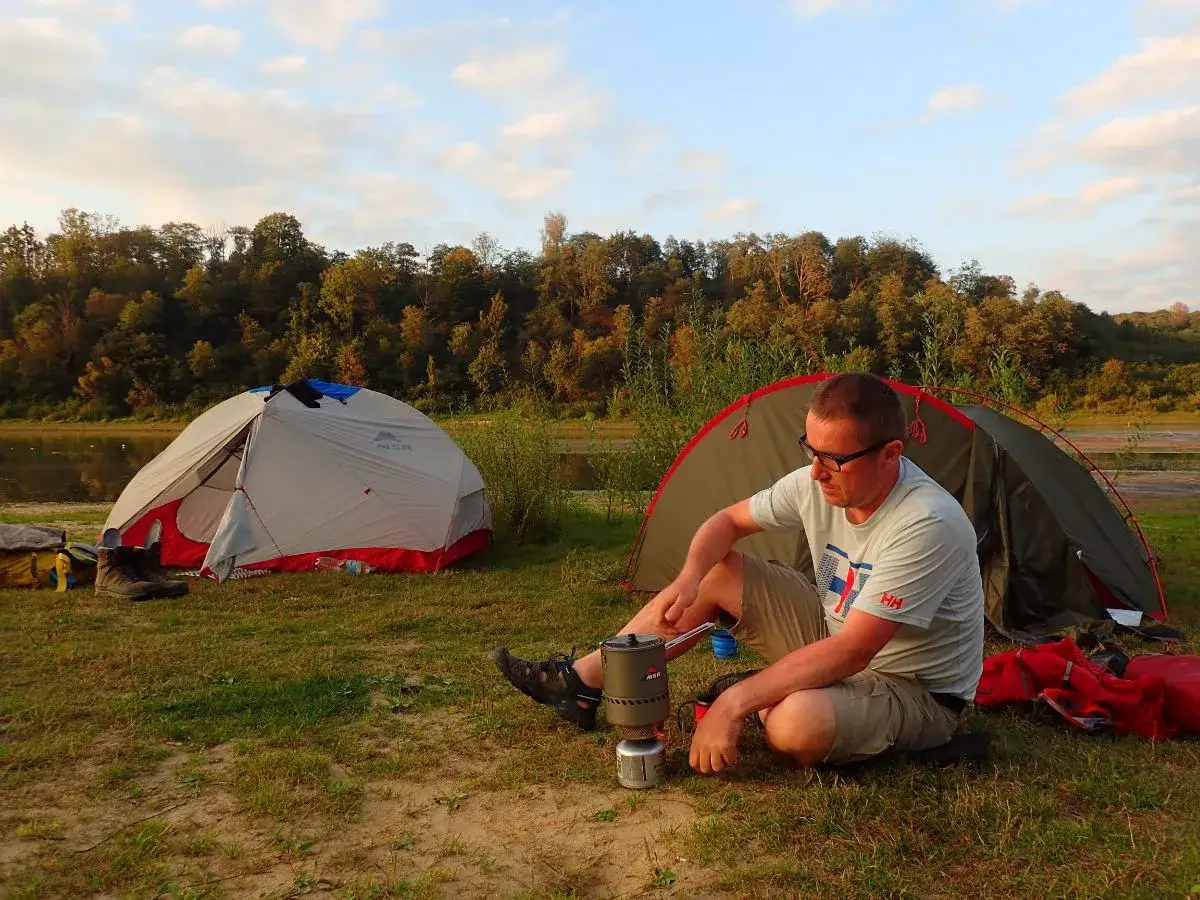 Mężczyzna siedzi przy rozłożonym namiocie, gotując coś na kuchence turystycznej. W tle drugi namiot i jezioro.