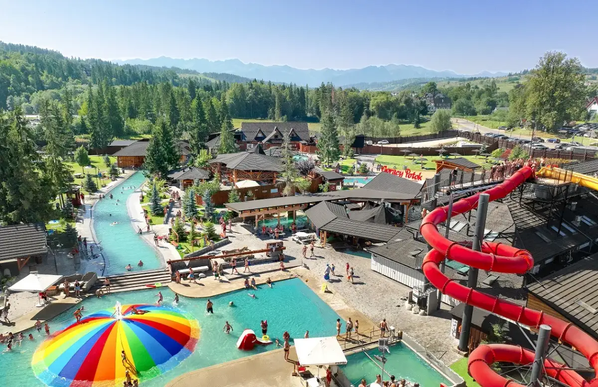 Widok z lotu ptaka na kompleks basenów termalnych z atrakcjami wodnymi, zjeżdżalniami i górami w tle.