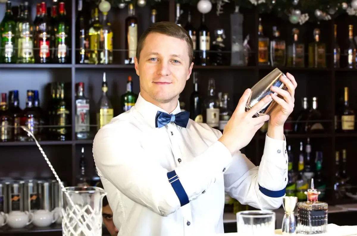 Uśmiechnięty barman w białej koszuli i niebieskiej muszce miesza drinka w shakerze na tle półek z butelkami alkoholu.