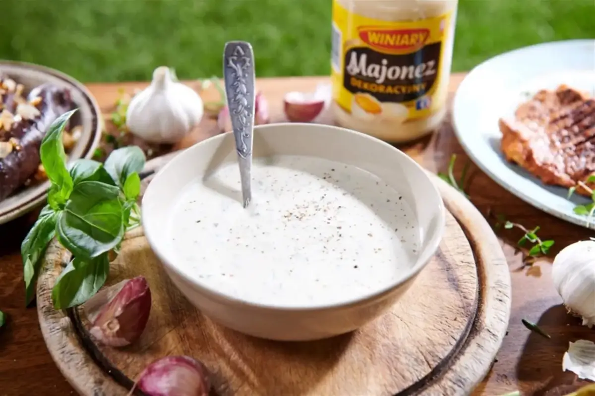 Miseczka z gęstym, białym sosem, łyżka, czosnek, zioła i słoik majonezu.