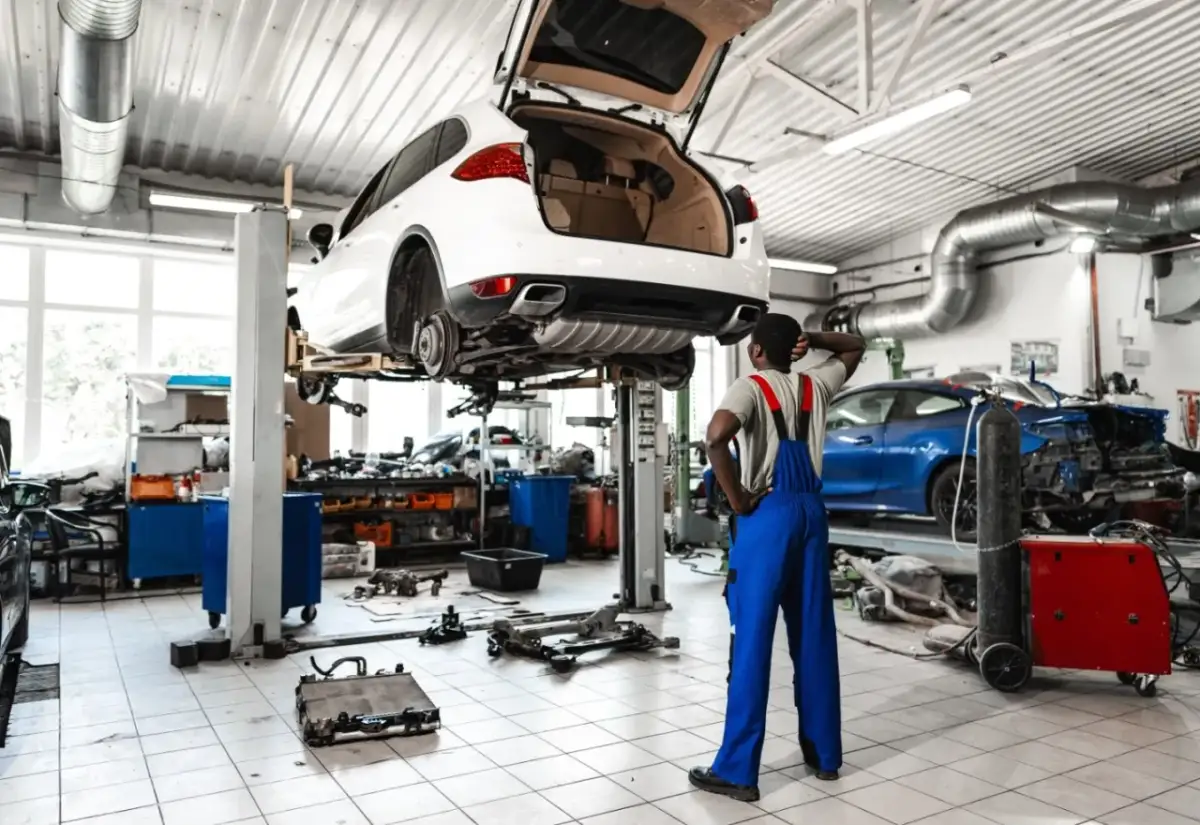 Mechanik zastanawia się, ile kosztuje założenie warsztatu samochodowego, patrząc na podniesione auto i części na podłodze.