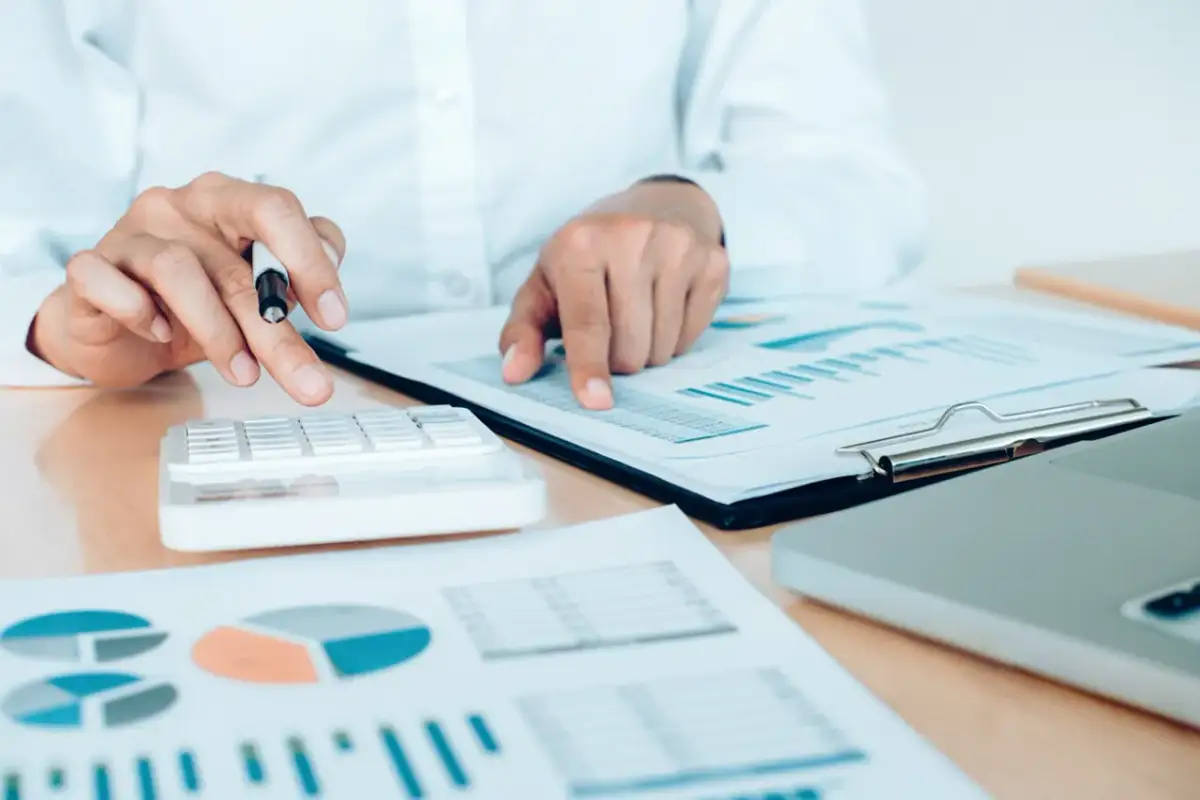 Dłonie osoby liczącej na kalkulatorze i wskazującej na wykresy i raporty.