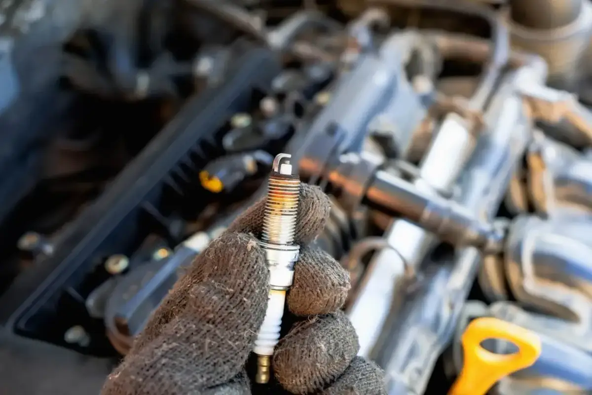 Ręka w rękawicy trzyma świecę zapłonową. W tle widać silnik samochodu. To może być objaw problemów z zapłonem.