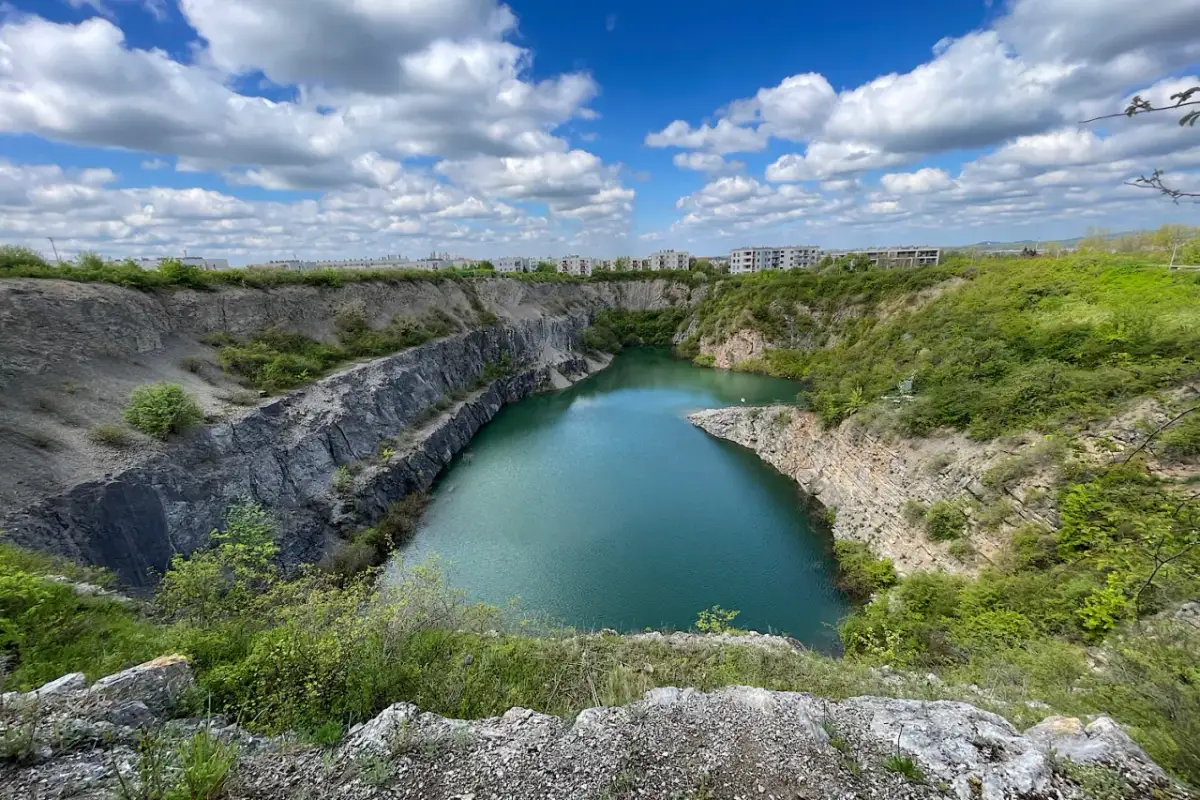 Wodny krajobraz w rezerwacie przyrody Ślichowice, z turkusową wodą w kamieniołomie otoczonym zielenią i budynkami w tle.
