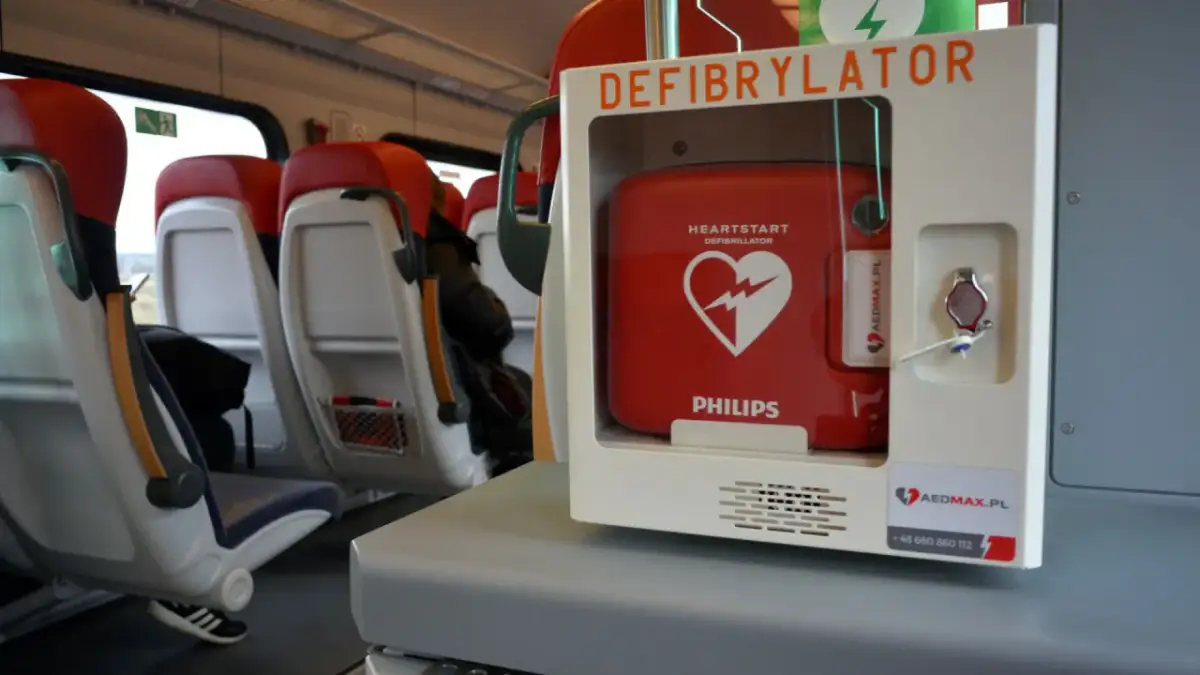 Czerwony defibrylator AED w białej obudowie, zamontowany w pociągu, obok siedzeń.