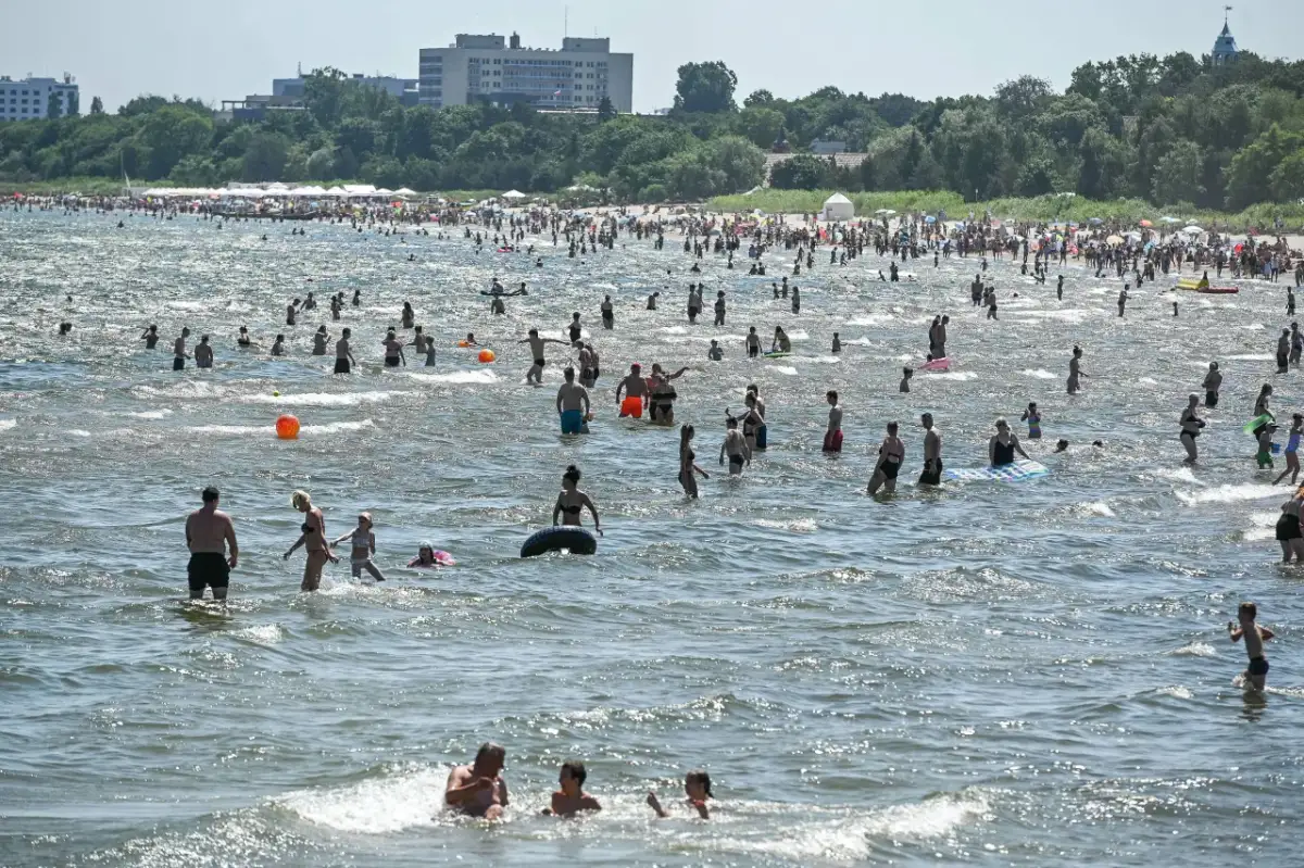 Tłum ludzi kąpiących się w Bałtyku, na tle plaży z parasolami i budynków.