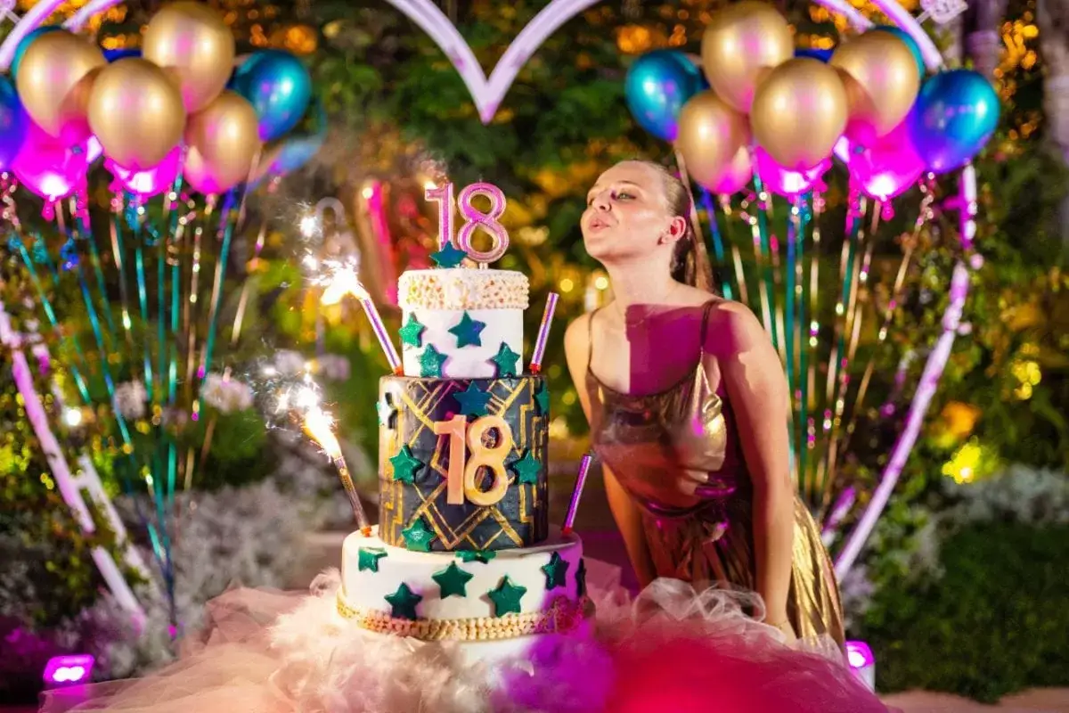 Dziewczyna w złotej sukience dmucha świeczki na torcie 18-stkowym, otoczona balonami i fajerwerkami.