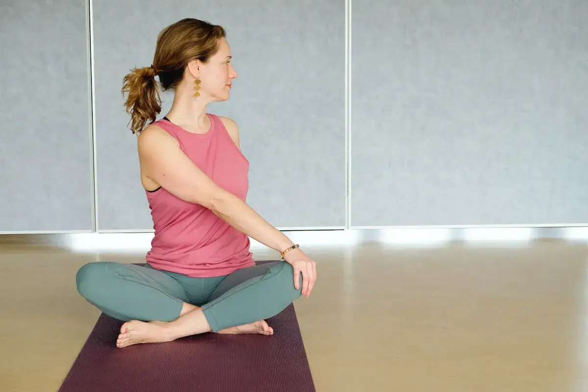 Kobieta w różowej bluzce i zielonych legginsach wykonuje skręty tułowia na macie do jogi.