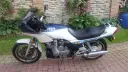 Klasyczny motocykl Yamaha XJ 900 w kolorze białym i granatowym, z niebieskimi i żółtymi pasami, zaparkowany na brukowanym podjeździe.