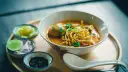 Aromaticzne danie z makaronem, które może być ramen lub udon, podane z dodatkami: limonką, cebulą i pikantnym sosem.