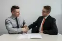 Dwóch mężczyzn w garniturach ściska sobie dłonie nad stołem, na którym leży dokument. To symboliczne podpisanie umowy przedwstępnej.