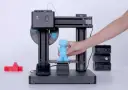Drukarka 3D z niebieską figurką i ręką wyjmującą ją z platformy.