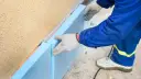 Ręce w rękawiczkach układają niebieski styropian przy ocieplanej ścianie fundamentowej.