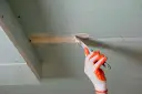 Ręka w rękawicy nakłada masę szpachlową na połączenie płyt gipsowo-kartonowych, przygotowując je do dalszych prac. Klejenie płyt GK na ściany wymaga precyzji.