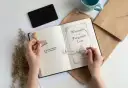 Ręce trzymają stempel z napisem "Biblioteczka Aleksandry" i książkę "Whispers of the Forgotten Lore". To idealny zestaw do tworzenia własnego exlibrisu.