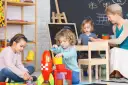 Dzieci bawią się klockami w żłobku. Nauczycielka nadzoruje zabawę, zastanawiając się, ile zarabia pomoc w żłobku.