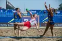 Piłkarka w locie rzuca zieloną piłką podczas meczu beach handball. Rywalki próbują ją zatrzymać.