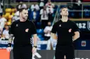 Dwóch sędziów w czarnych koszulkach z logo Hummel i emblematem związku sportowego, gotowych do rozpoczęcia meczu ligi piłki ręcznej.