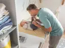 Ojciec całuje stópki dziecka na przewijaku. Wymiary przewijaka są idealne do pielęgnacji malucha. Obok stosik ubranek.