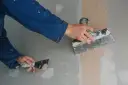 Ręce w niebieskim kombinezonie wykonują szpachlowanie ścian, wygładzając masę szpachlową za pomocą narzędzi.