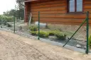 Montaż siatki ogrodzeniowej przy drewnianym domu. Widać zielone słupki, siatkę i betonowy podmurówkę.