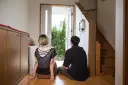 Para siedzi na podłodze w sieni, patrząc na otwarte drzwi. Widok na zielone drzewa.
