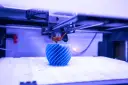 Drukarka 3D tworzy niebieski, spiralny obiekt na białej platformie.