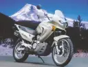 Srebrny motocykl Honda Transalp 650 gotowy na przygodę na tle ośnieżonej góry i lasu.