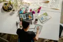 Kobieta pracuje online w domu, projektując na laptopie i tablecie. Na biurku kwiaty, kubek kawy i czaszki z pisakami.
