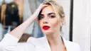 Frau mit roten Lippen und weißem Blazer, Hand am Kopf.