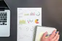 Laptop, arkusz z wykresami (liniowymi, słupkowymi, kołowymi) i notatnik z ręką trzymającą długopis.