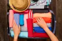 Ręce układają kolorowe ubrania w walizce. Słomkowy kapelusz i wzorzysty strój kąpielowy czekają na podróż. To gotowy bagaż na wakacje.