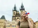 Uśmiechnięta blondynka z flagą Czech zasłaniającą oko, marzy o wysokich zarobkach w Czechach. W tle praskie kamienice i kościół.