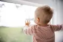 Małe dziecko w paski patrzy przez okno, dotykając szyby.