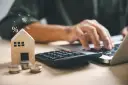 Drewniany domek, monety i kalkulator symbolizują planowanie finansowe. To idealny obraz dla osób szukających mieszkania na start.