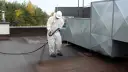 Pracownik w białym kombinezonie natryskuje dachową papę, zabezpieczając ją przed wilgocią.