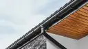 Czarny system rynnowy odprowadza wodę opadową z dachu. Widoczny fragment elewacji z kamienia i drewnianego podsufitki.