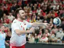 Piłkarz ręczny Polski w akcji, rzuca piłkę. W tle widać trybuny z kibicami, którzy dopingują swoją drużynę w meczu przeciwko Iranowi.