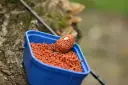 Zbliżenie na niebieskie pudełko z pomarańczowym pelletem, idealnym jako jaki pellet do metody na karpia. Na wierzchu znajduje się podajnik z przynętą.