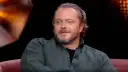 Tomasz Schuchardt, znany z wielu filmów, seriali i programów, z charakterystyczną fryzurą i niebieskimi oczami, siedzi na kanapie.