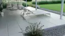 Nowoczesny taras betonowy z leżakami i sofą, otoczony trawą i kamieniami. Idealne miejsce na relaks.