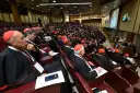 Wielu kardynałów w czerwonych biretach i czarnych szatach siedzi w amfiteatrze, słuchając przemówienia podczas ważnego konsystorza.