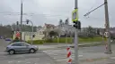 Szare auto zbliża się do przejścia dla pieszych, gdzie światła kolizyjne regulują ruch. W tle budynki i drzewa.