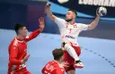 Gracz w piłkę ręczną w skoku, z piłką w dłoni, celuje do bramki. Jego przeciwnik próbuje go zatrzymać.