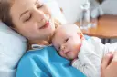 Uśmiechnięta mama tuli noworodka. To chwila spokoju po szczepieniach noworodka, budująca więź i zapewniająca zdrowie maluszka.